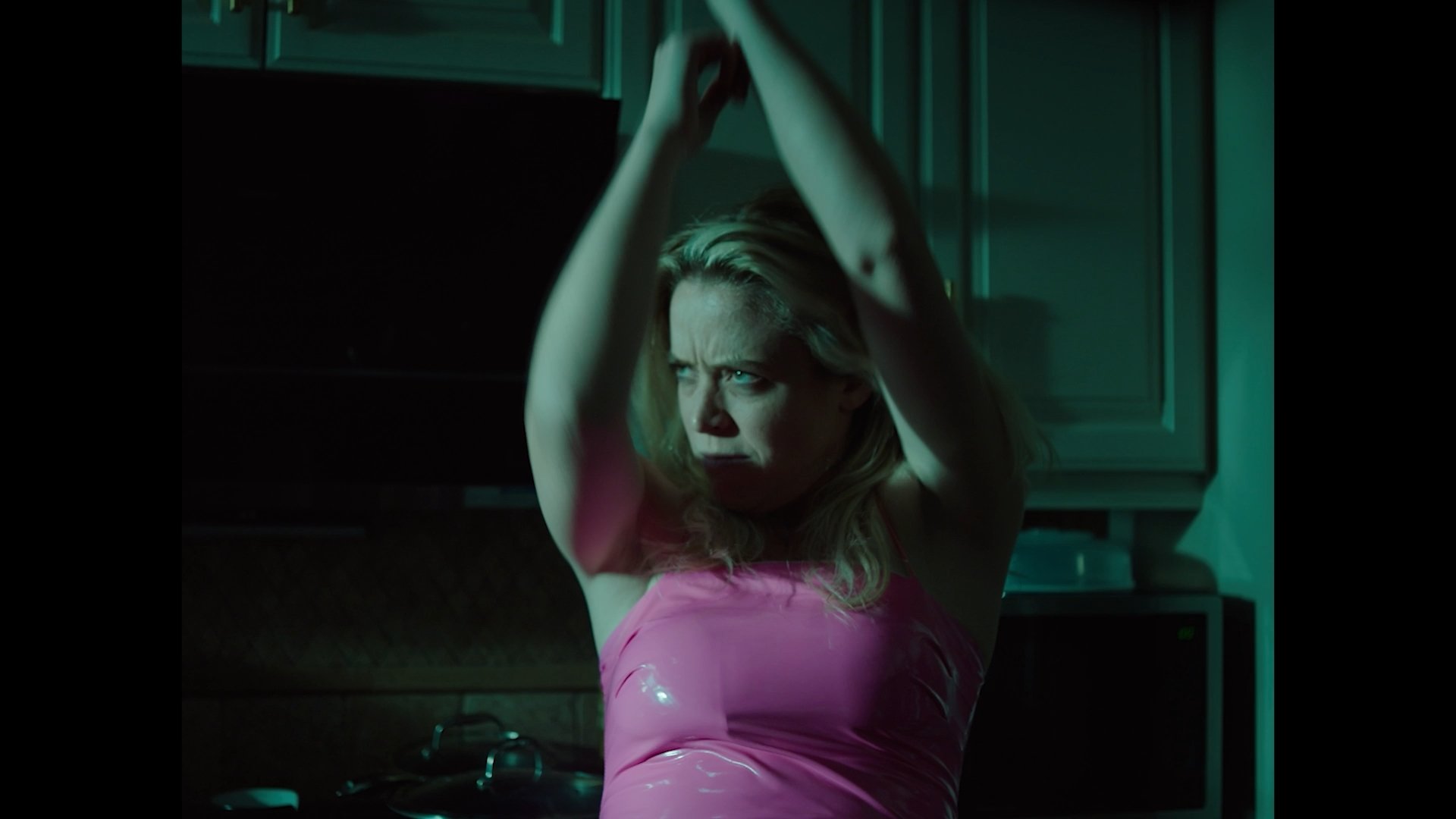 A woman with blonde hair wearing a pink top, standing with her arms raised above her head in a room with dark and greenish lighting.