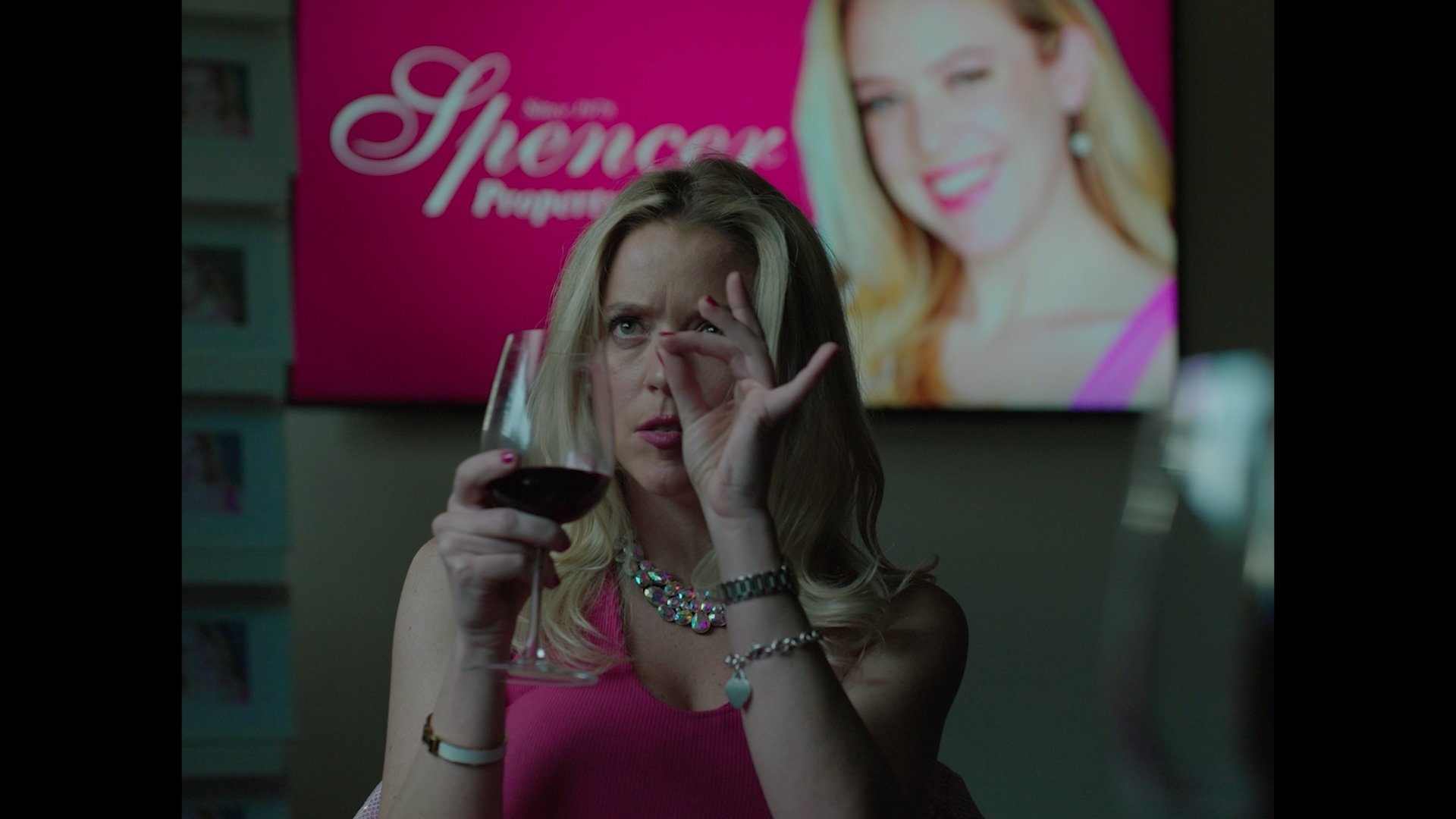 A woman with blonde hair in a pink top is holding a glass of red wine, looking through her fingers with a playful expression. Behind her, there is a bright pink advertisement poster featuring a woman with red hair.