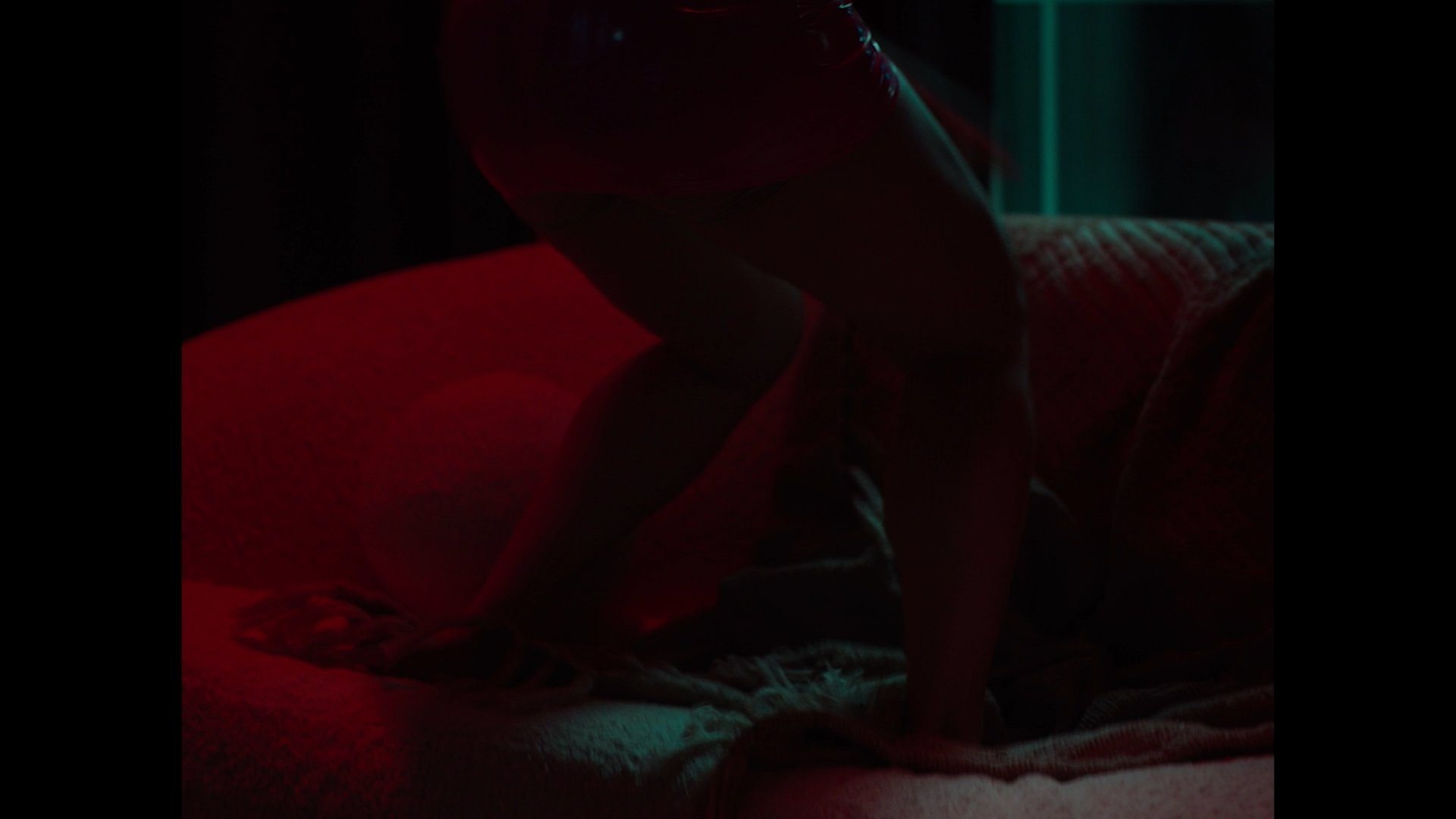 A person is on a bed in a dark room illuminated by red and teal lighting, with their body bent forward and arms on the bed.