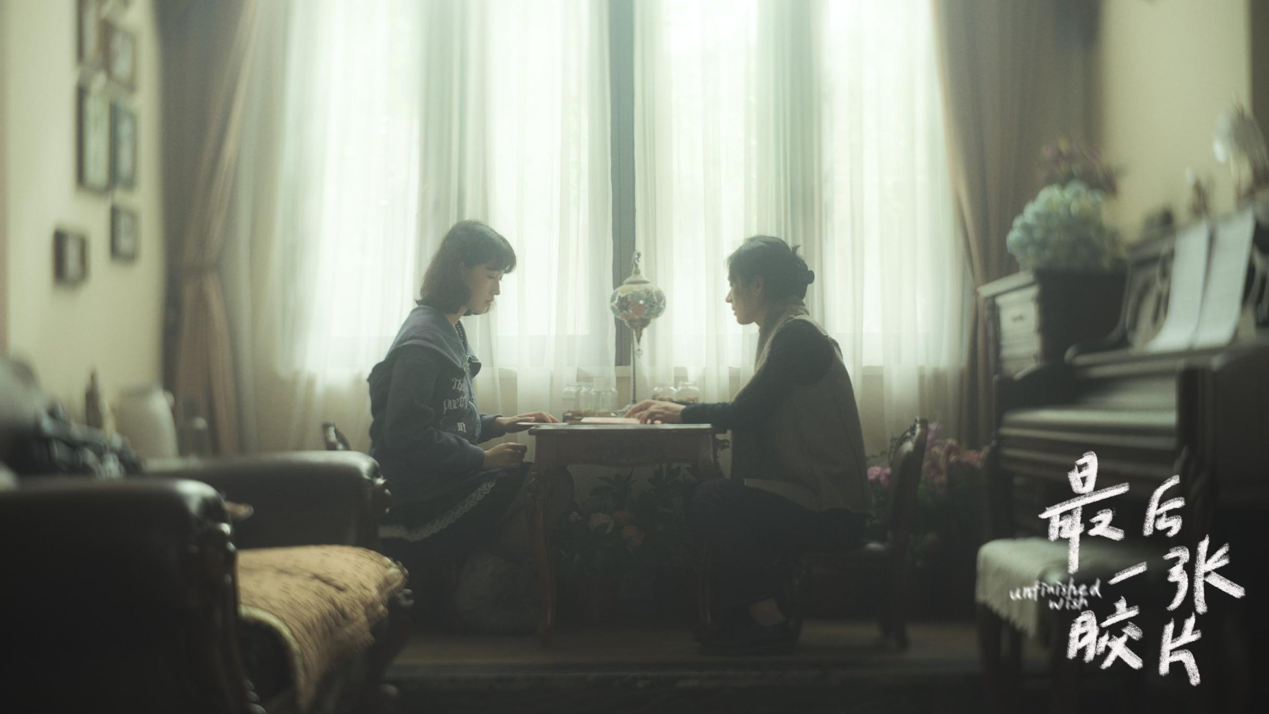 Two women sit across from each other at a small table in a well-lit room with large windows and white curtains, engaging in conversation.