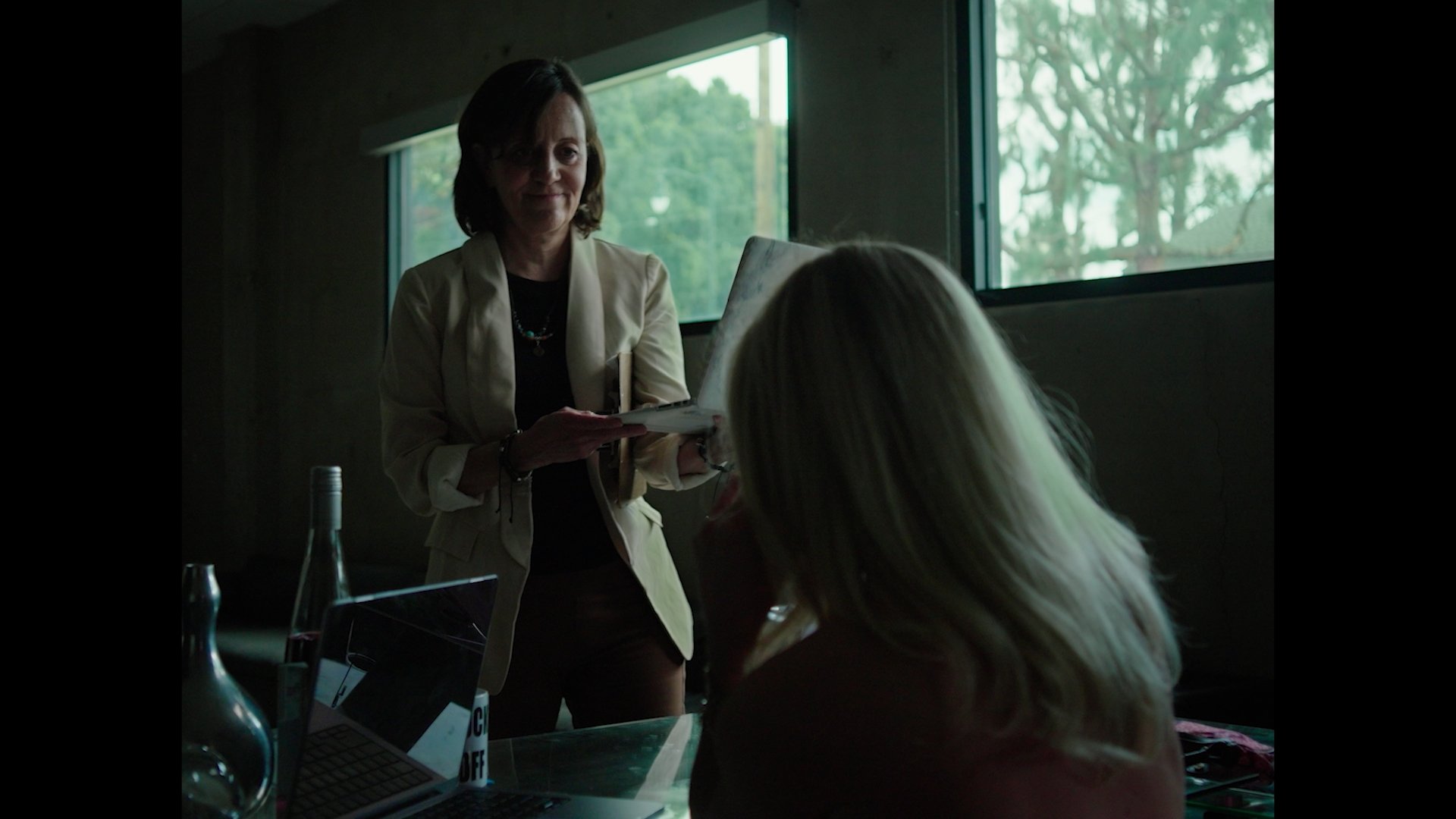 A woman with short dark hair standing and smiling while holding a tablet, facing a seated person with long blonde hair in a room with large windows showing trees outside.