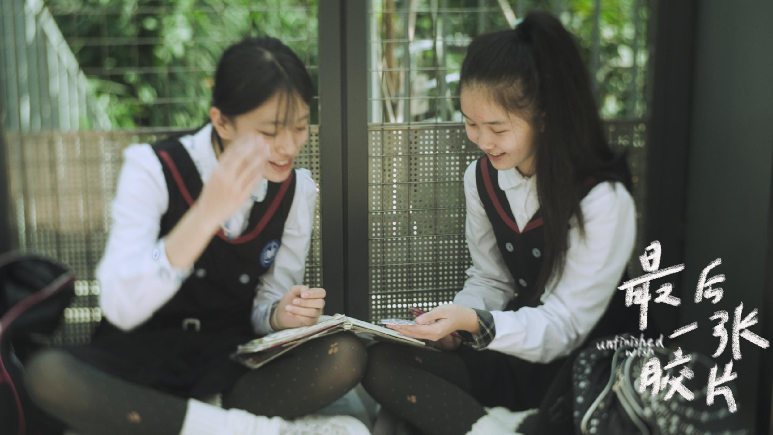 Two schoolgirls in uniforms sitting on the ground, looking at a booklet and smiling, with a metal fence and greenery in the background.