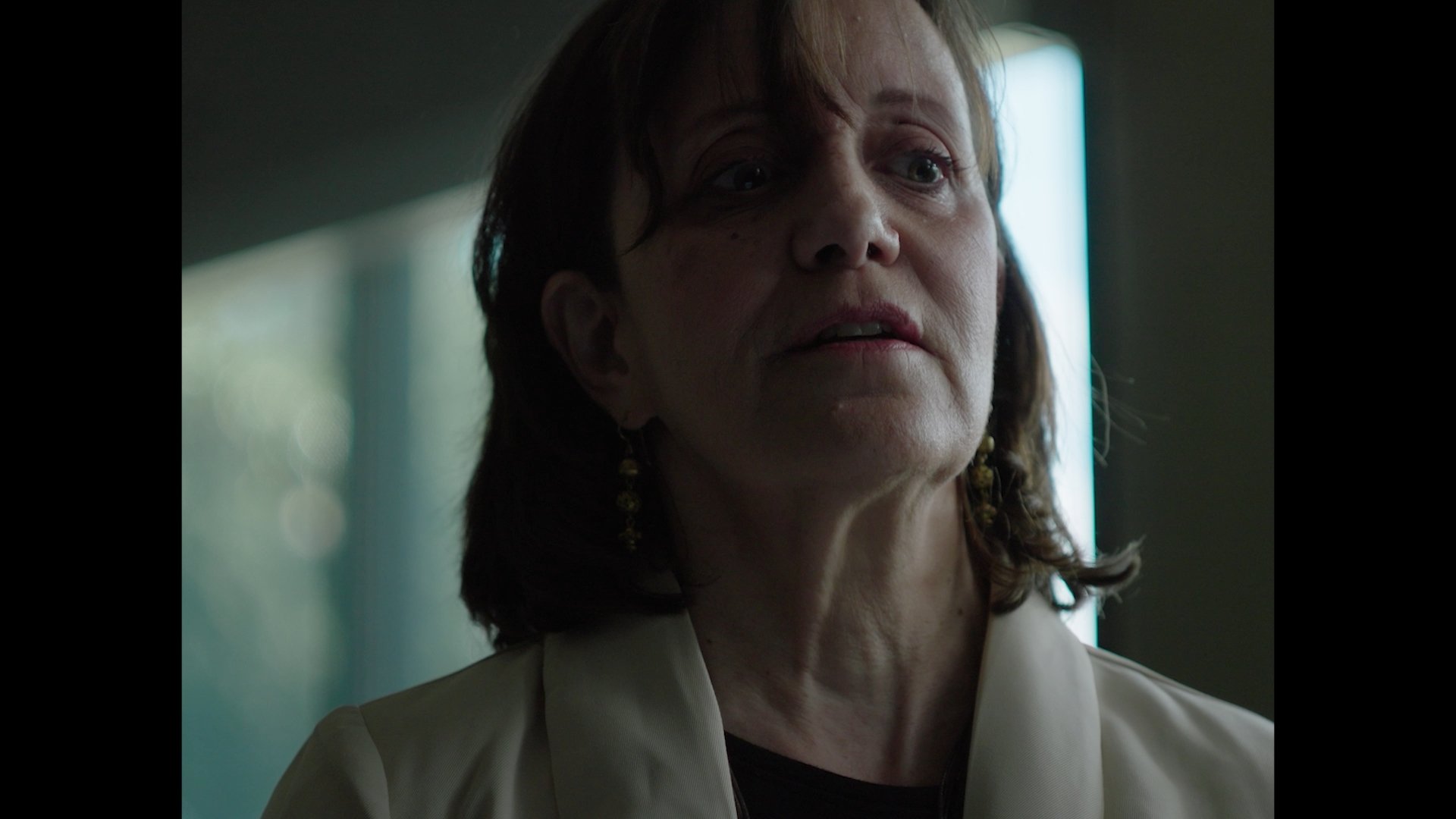 Close-up of a middle-aged woman with reddish-brown hair, wearing a beige blazer and earrings, looking slightly to the side in a dimly lit room with a bright vertical light in the background.